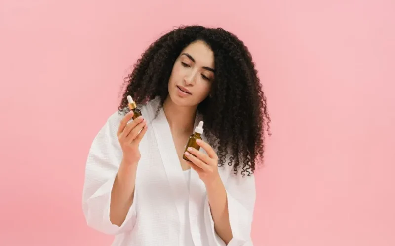 A woman with curly hair in a white robe examines two cosmetic oil bottles against a pink background.