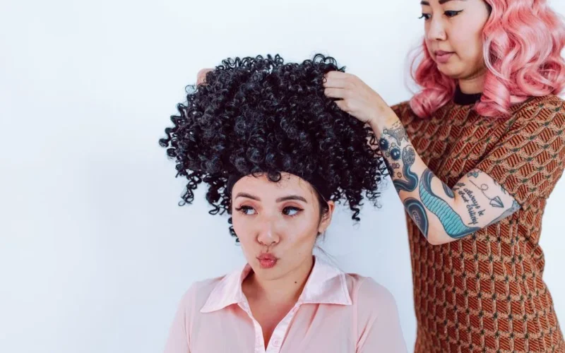 A hairstylist with tattoos fits a curly black wig on a woman in a salon.