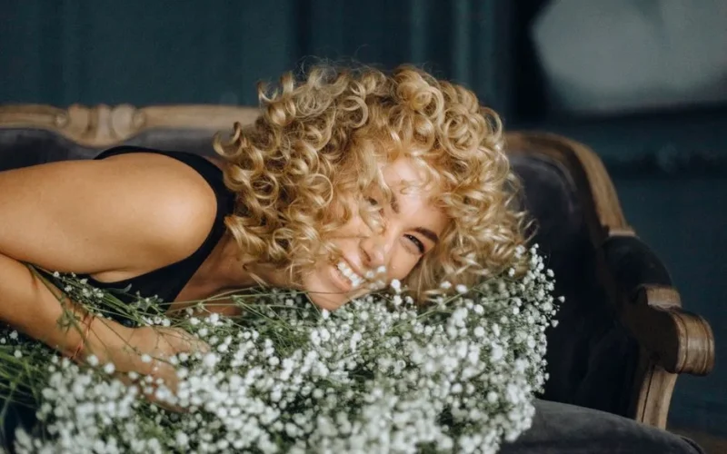 Smiling woman with curly blond hair resting on a couch with white flowers indoors.