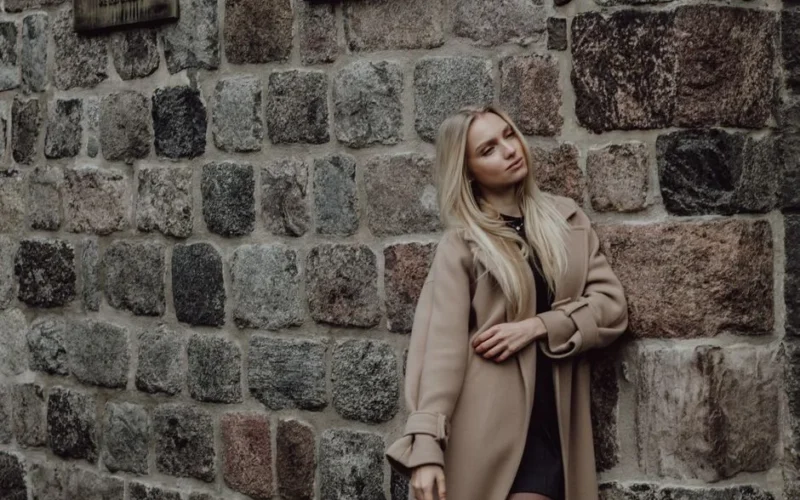 A fashionable woman with blonde hair in a beige coat leans against a rustic stone wall.