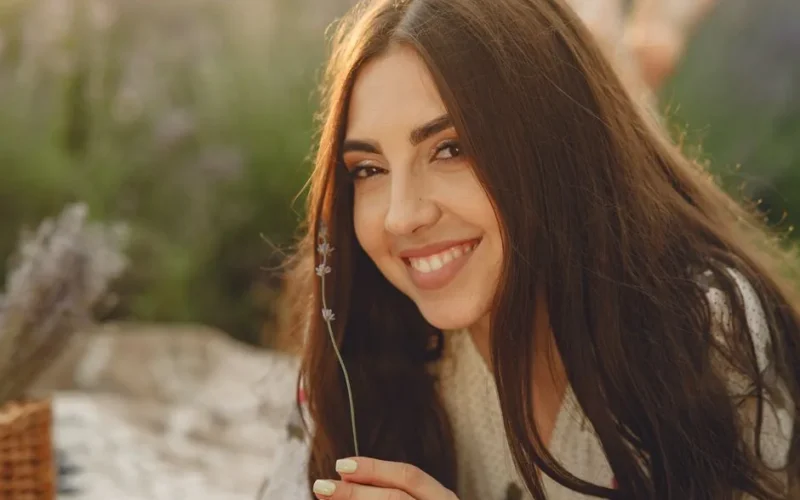 A happy woman with long brunette hair holds a flower outdoors.