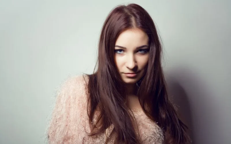 Close-up portrait of a young woman with long brown hair and a stylish outfit against a neutral background.