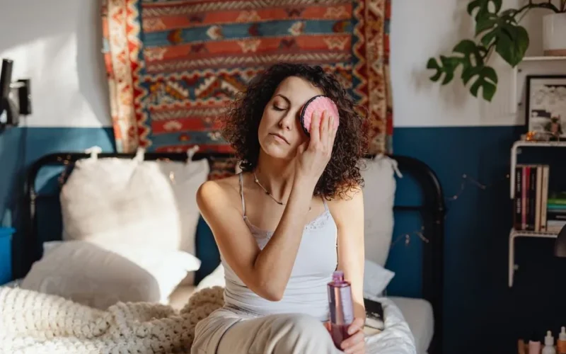Woman with curly hair removes makeup using a cotton pad in a cozy, stylish bedroom.