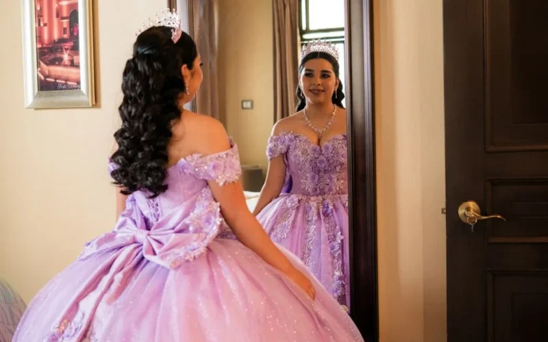 Elegant young woman in a lavender gown and tiara admiring her reflection in an ornate indoor setting.