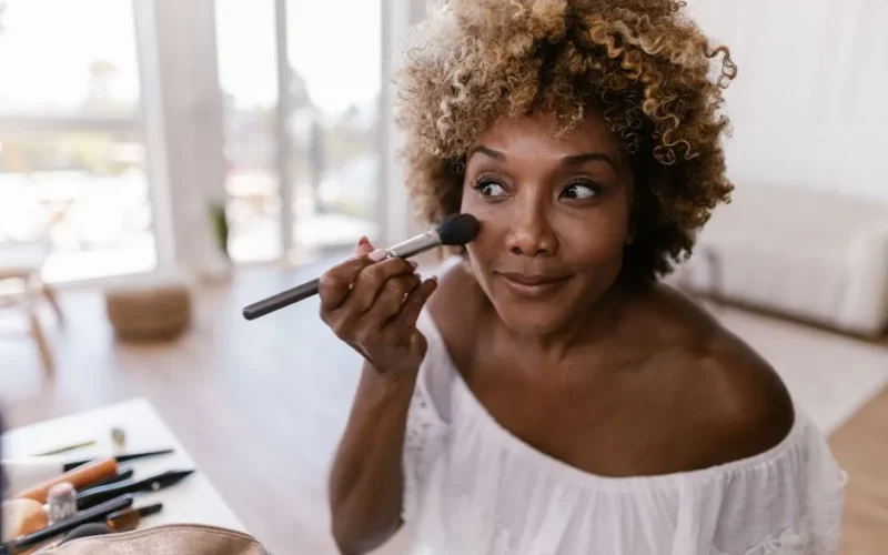 Woman with curly hair applying makeup inside a well-lit room, creating a natural look.