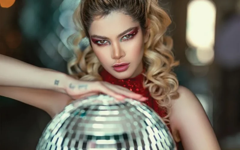 Stylish woman with expressive eyes poses with a disco ball, glamorous makeup enhancing her features.