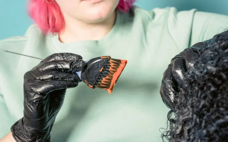 Crop female hairdresser in black gloves applying bright pigment on curly hair of client