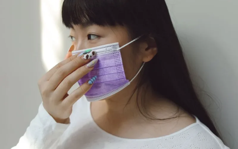 Asian woman wearing a purple medical mask, practicing personal safety indoors with brightly polished nails.