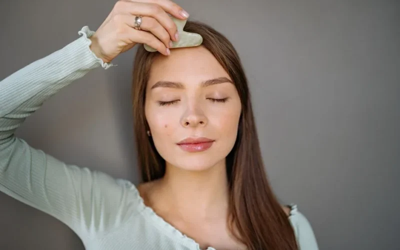 A woman relaxes with a jade gua sha tool for a facial skincare routine against a gray background.