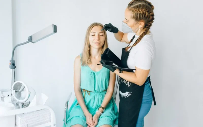 A beautician performs eyebrow microblading on a woman in a modern salon setting.