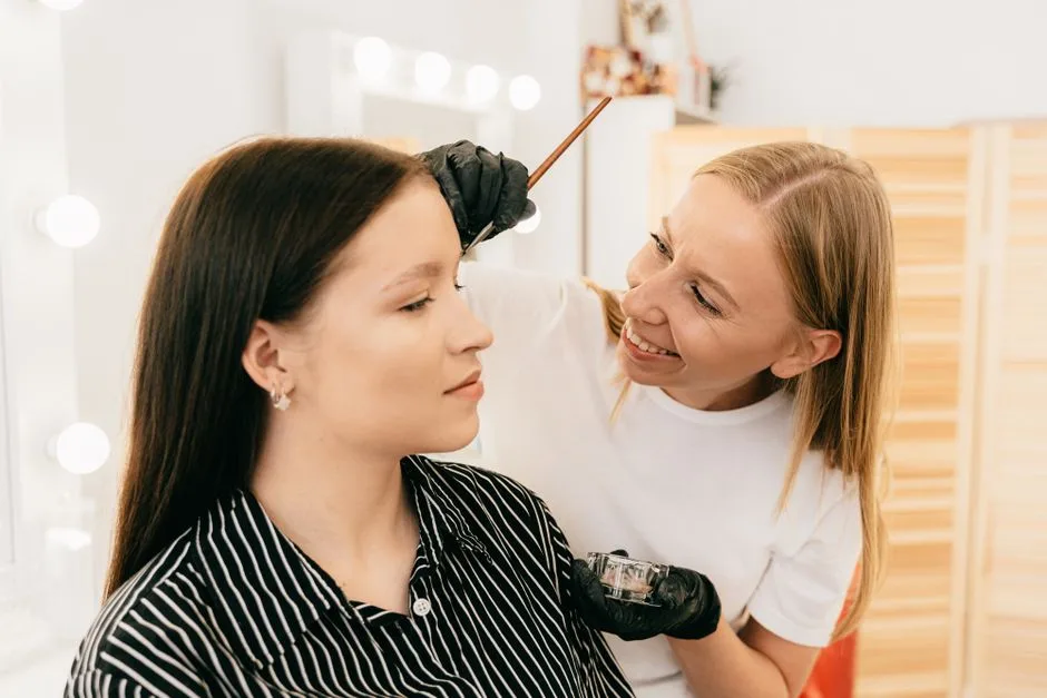Makeup artist applying eyebrow tint on a smiling woman in a beauty salon.