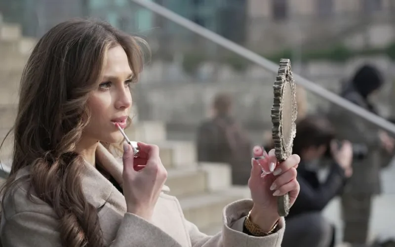 A fashionable woman applies lipstick while gazing into a hand mirror on a Berlin street.