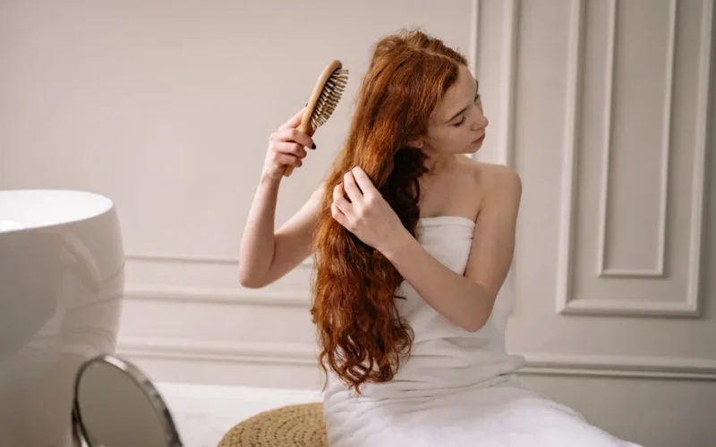 Woman with long red hair wrapped in a towel combing her hair in an elegant bathroom setting.