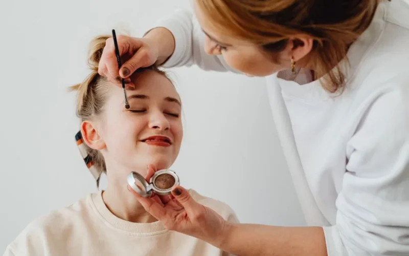 Makeup artist applying eye shadow to a smiling teen girl. Close up shot highlighting beauty and cosmetics.
