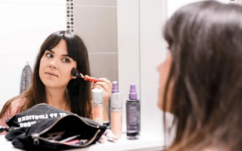 A woman applying makeup using a brush in front of a bathroom mirror, surrounded by cosmetics.