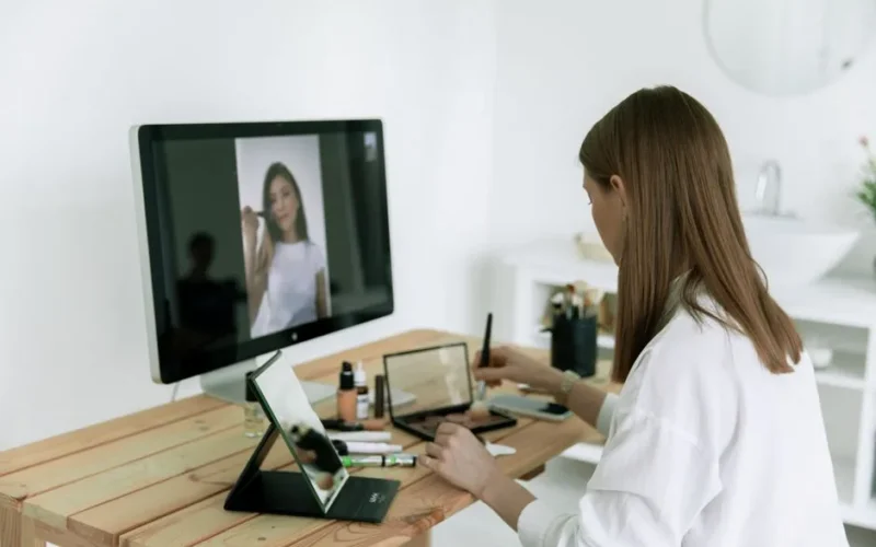A woman follows an online makeup tutorial using a computer and tablet at home.