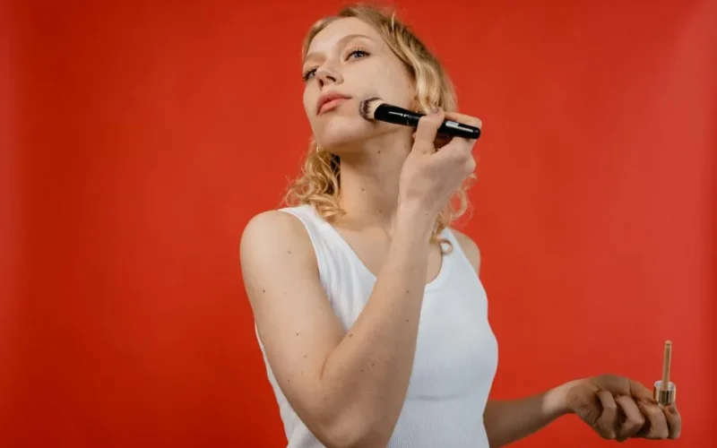 Woman uses a makeup brush to apply cosmetics in front of a vivid red background.