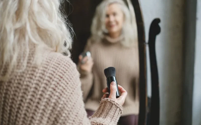 A senior woman applies makeup while looking in a mirror, holding a brush in her hand.