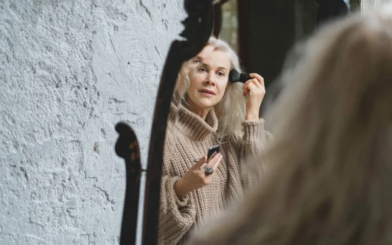 Elderly woman puts on makeup while looking into a mirror at home.