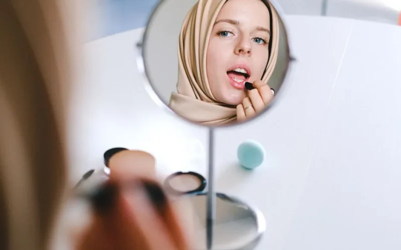 Muslim woman in hijab applies lipstick while looking at her reflection in a mirror.