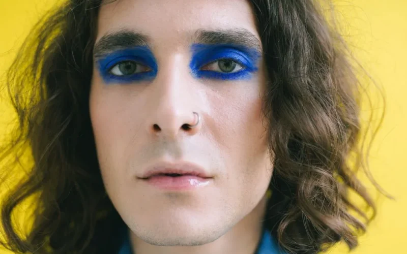 Close-up portrait of a person with striking blue eye makeup against a yellow backdrop.
