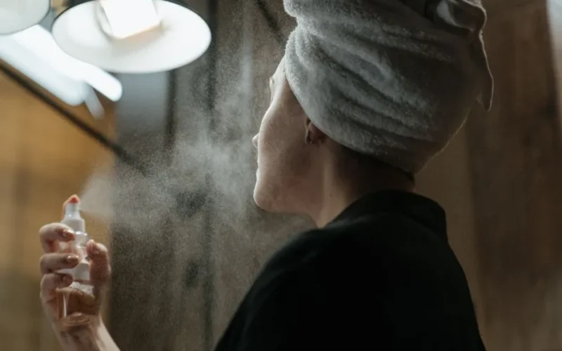 Woman with a towel on head applying facial spray in bathroom, focusing on skincare and self-care.