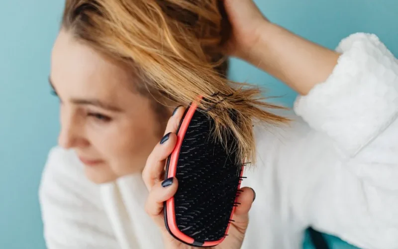 Close-up of a woman brushing her blonde hair with a red hairbrush indoors.