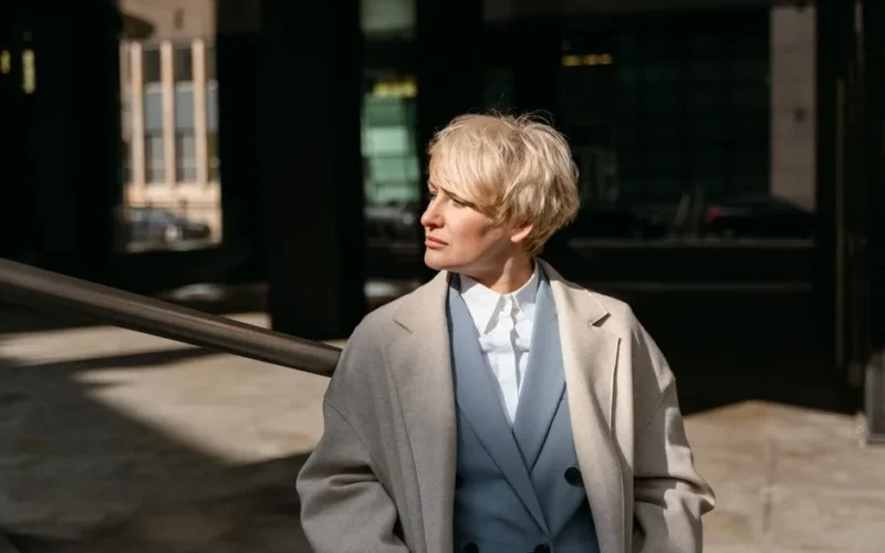Professional blonde woman in a coat, facing away in a sunlit urban environment.