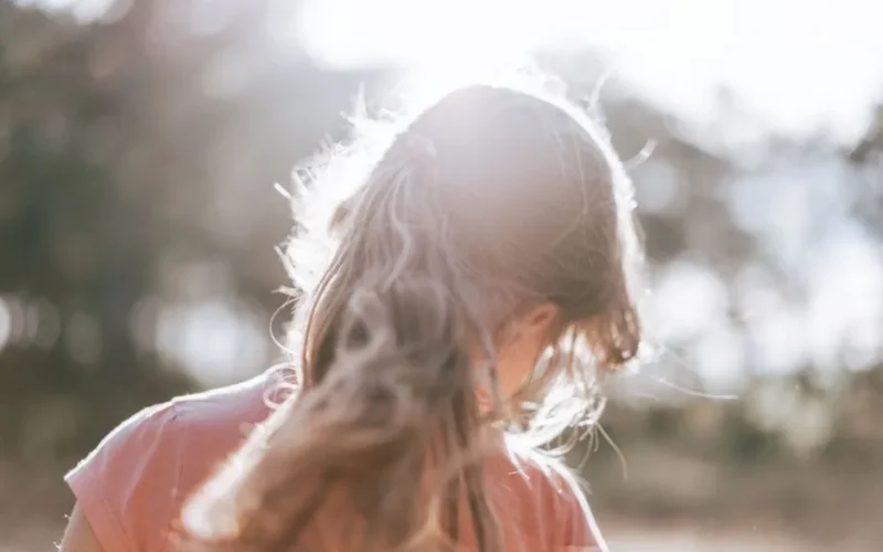 Back view of blond haired girl with ponytail in casual clothes standing on meadow near forest on sunny summer day