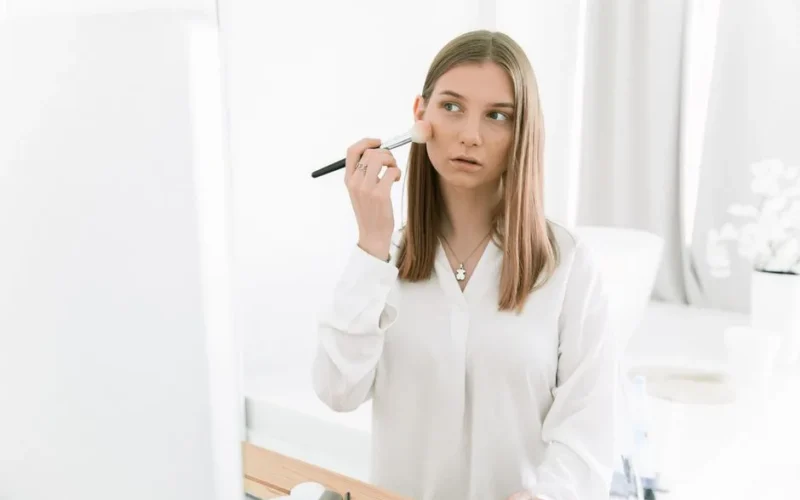 A woman wearing white long sleeves applies blush indoors using a makeup brush.