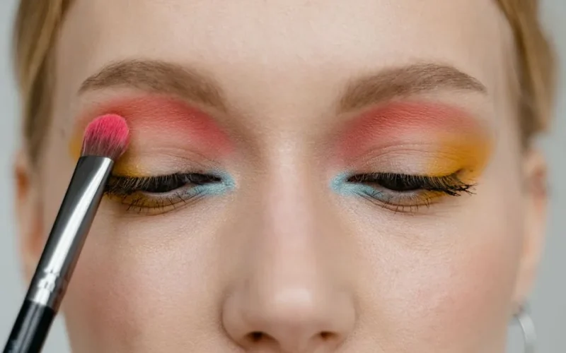 Vibrant eye makeup close-up with brush applying pink, blue, and yellow colors.