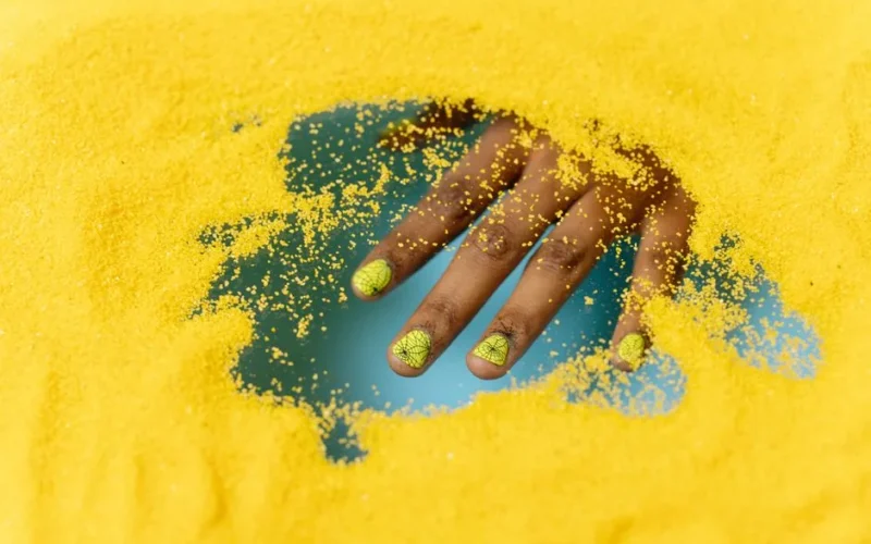 Close-up of a hand with creative nail art partially buried in vibrant yellow sand.