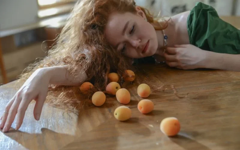 A woman lies pensively with apricots on a wooden table, creating a reflective mood.