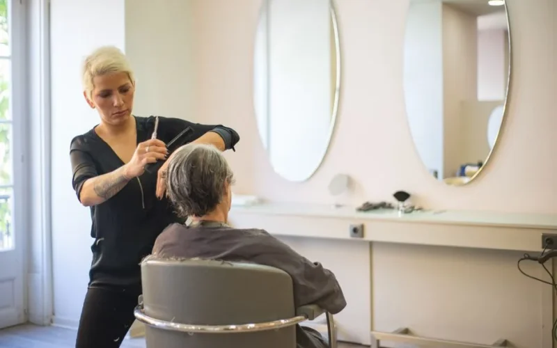 A hairdresser skillfully cuts an elderly woman's hair in a modern salon with mirrors and a comfortable barber chair.