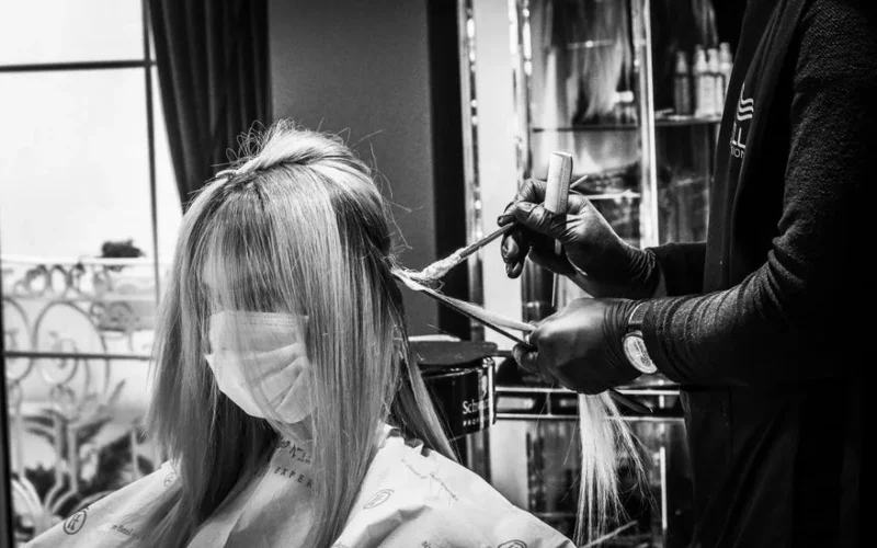 Hair stylist applying color to a woman wearing a mask in a salon, in black and white.
