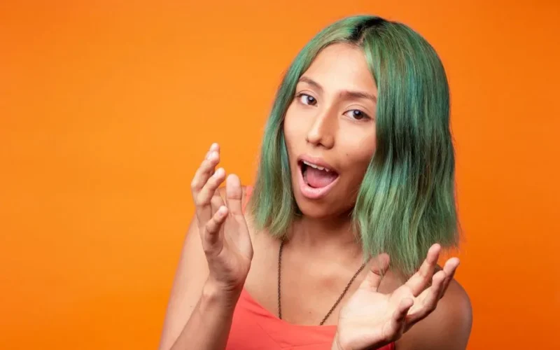 Portrait of a young woman with green hair against an orange background, expressing emotion.