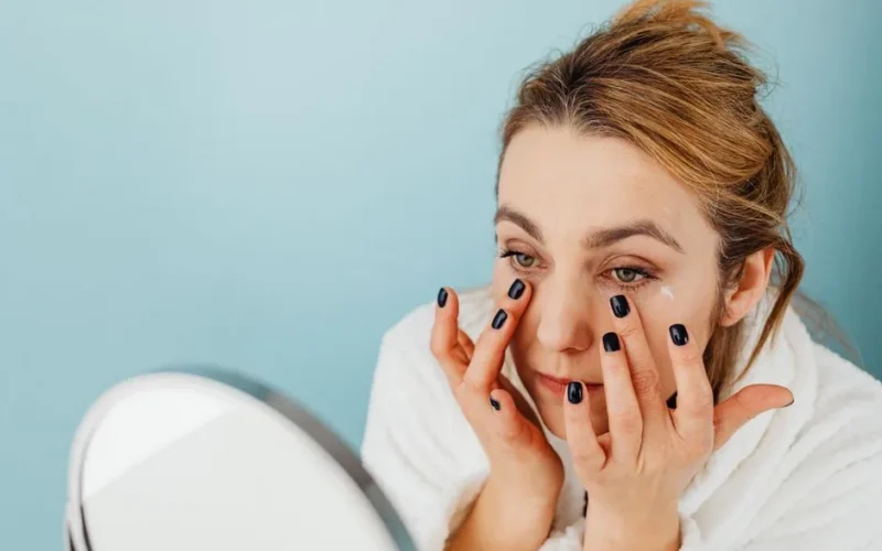 Woman applies eye cream on her under-eye area with black nails, wearing a white robe.