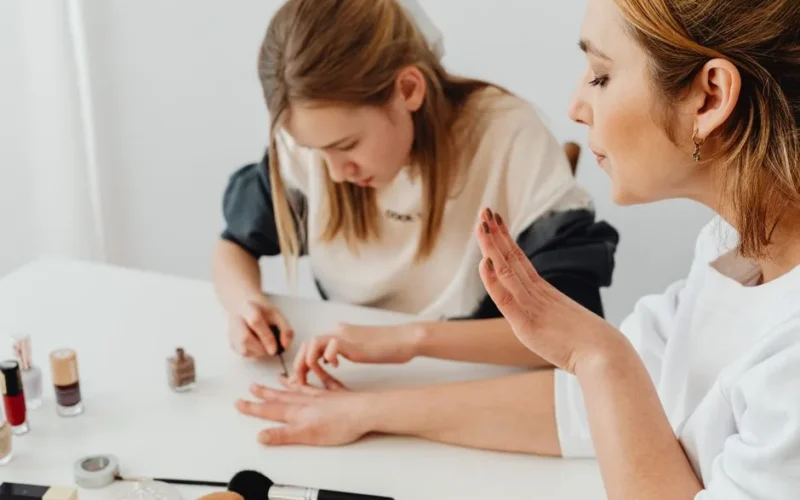 A young girl paints a woman's nails during a home manicure session at a table.