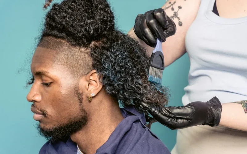 Crop female hairdresser in gloves with brush spreading dye on hair of African American male customer sitting against blue background