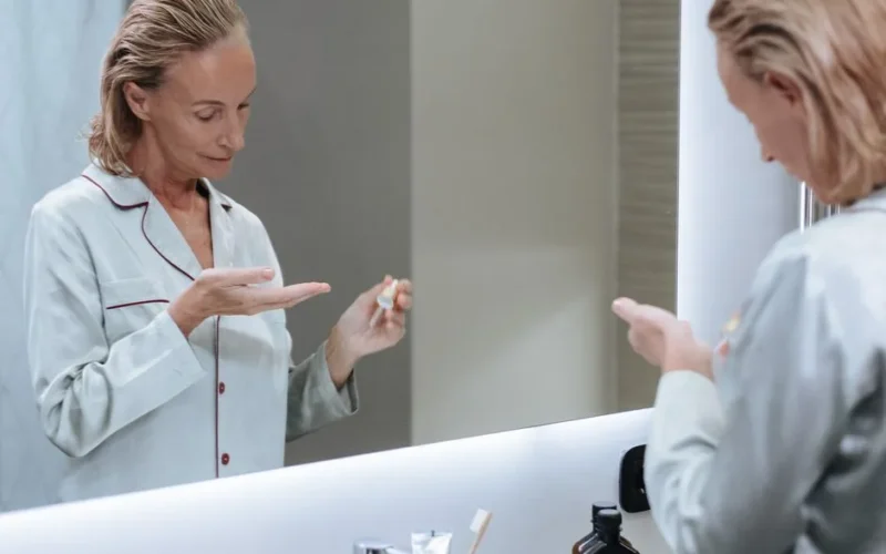 Senior woman applying skincare serum in front of bathroom mirror during her morning routine.