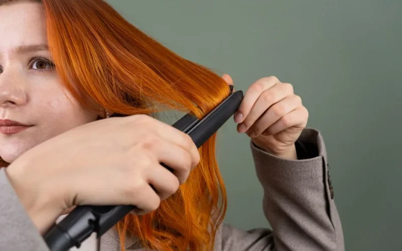 Close-up of a woman styling her red hair using a hair straightener against a green background.