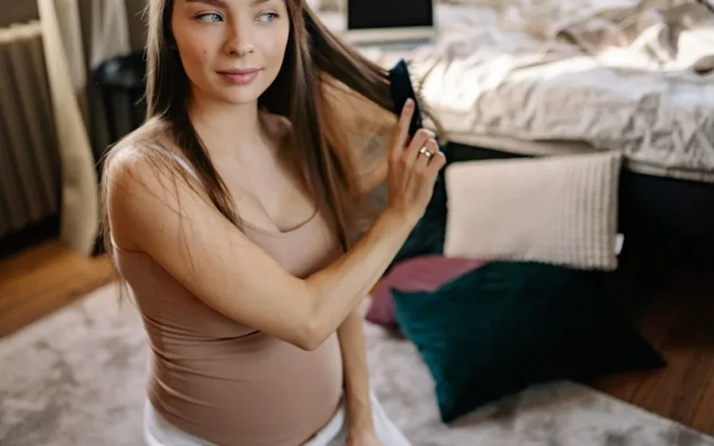A pregnant woman combing her hair while sitting in a cozy bedroom environment.