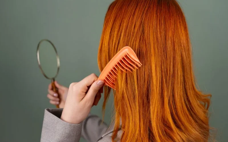 Close-up of a woman with red hair combing while holding a mirror.