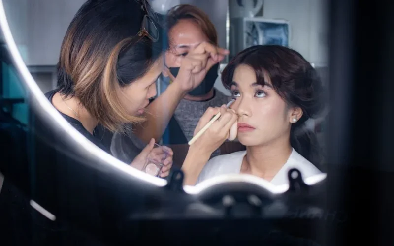 A makeup artist applies eye makeup to a bride indoors, captured in a professional setting.
