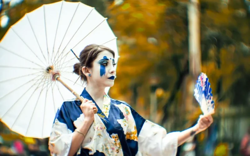 A woman in a blue kimono with dramatic makeup holds a parasol in an outdoor setting, capturing cultural elegance.