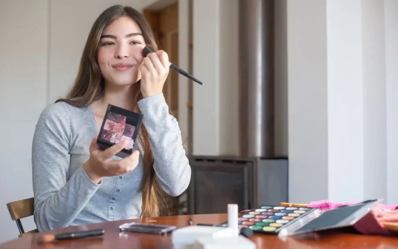 Young woman applying blush on cheeks with a makeup brush at home, smiling.