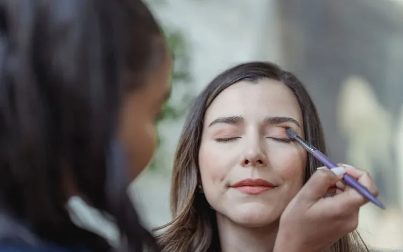 Crop young ethnic female makeup artist using brush while applying eyeshadow on eyelid of client with closed eyes in light beauty salon