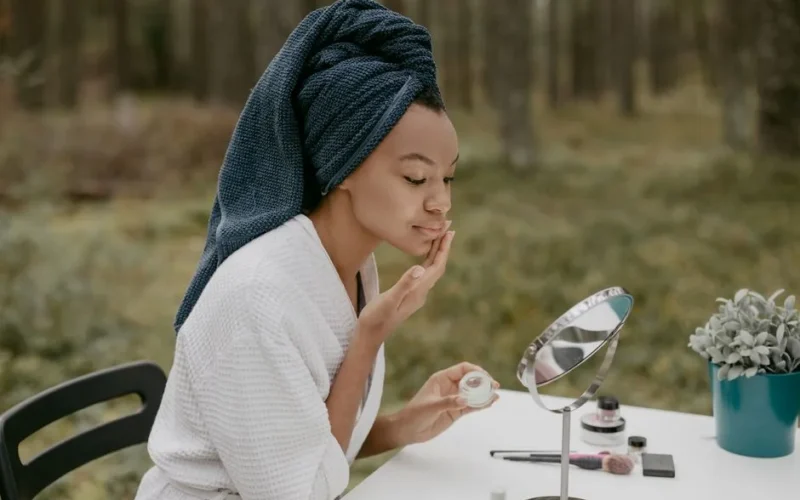A woman in a bathrobe applies face cream in a serene outdoor environment.