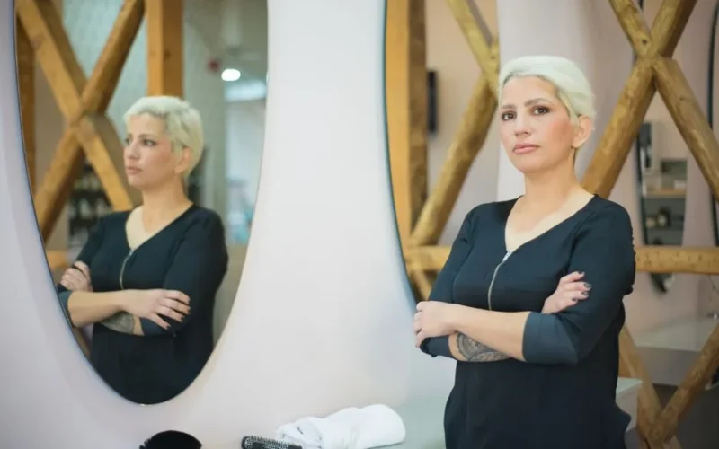 A professional hairdresser confidently stands in a modern salon with crossed arms, reflecting in a mirror.
