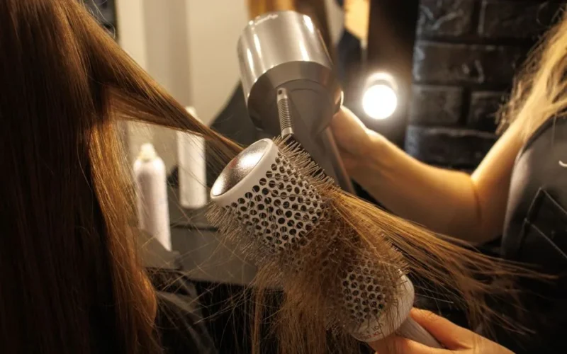 Close-up of a hairstylist drying and styling long hair with a round brush in a salon.